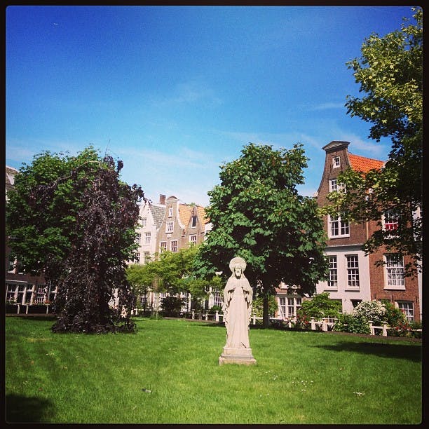 En gräsbevuxen innergård med en staty av en person nära centrum, omgiven av träd och traditionella byggnader i europeisk stil.