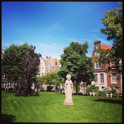 En gräsbevuxen innergård med en staty av en person nära centrum, omgiven av träd och traditionella byggnader i europeisk stil.