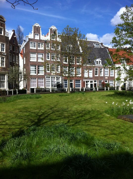 Välskött gräsmatta och några träd framför traditionella tegelbyggnader med vitt inramade fönster under en blå, delvis molnig himmel.