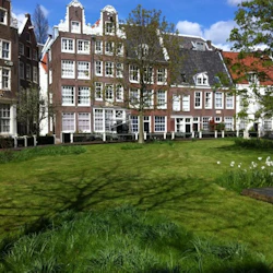 Välskött gräsmatta och några träd framför traditionella tegelbyggnader med vitt inramade fönster under en blå, delvis molnig himmel.