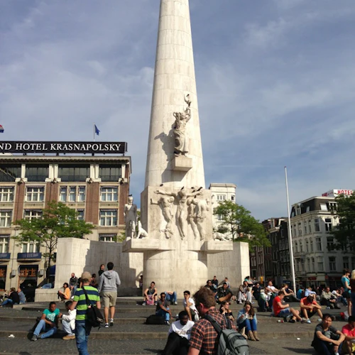 Människor som sitter och går framför ett högt obeliskmonument med skulpturer, på ett torg nära byggnader och träd.