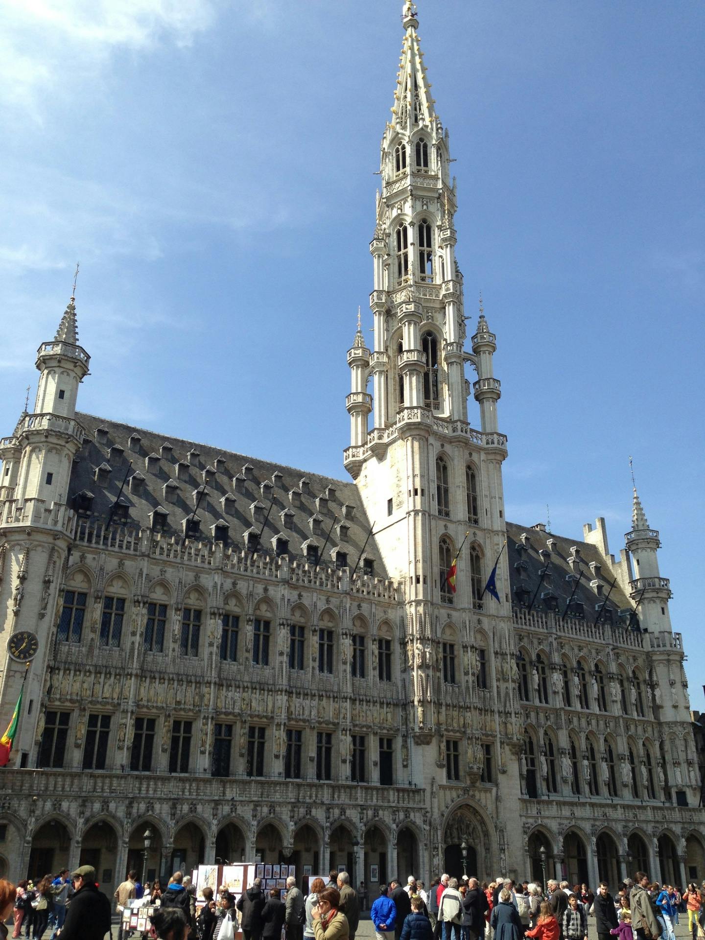 Stor historisk byggnad med en hög utsmyckad spiral, grått tak och välvda fönster. Människor samlas framför en solig dag.