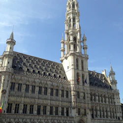 Stor historisk byggnad med en hög utsmyckad spiral, grått tak och välvda fönster. Människor samlas framför en solig dag.