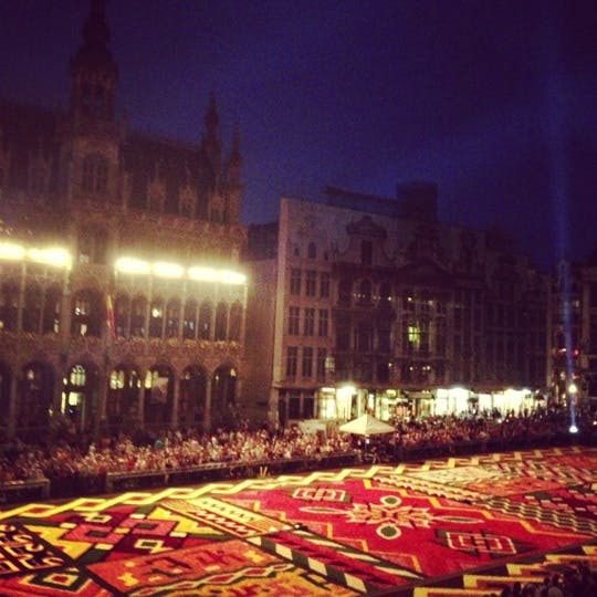 En stor, färgglad blommig matta visas på ett torg på natten, med historiska byggnader och en folkmassa i bakgrunden.