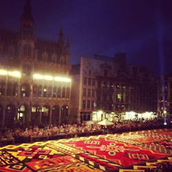 En stor, färgglad blommig matta visas på ett torg på natten, med historiska byggnader och en folkmassa i bakgrunden.