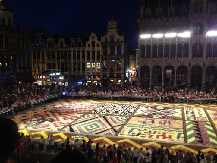 En stor, färgstark blommig uppvisning på ett torg, omgiven av historiska byggnader och en folkmassa som observerar på natten.