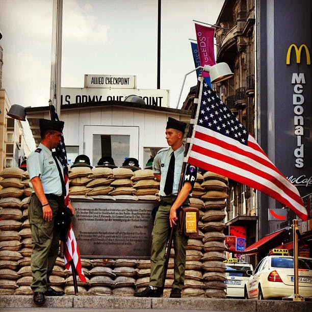 Dois guardas com uniformes militares estão no Checkpoint Charlie em frente a sacos de areia, segurando bandeiras americanas, com edifícios e um sinal do McDonald's ao fundo.