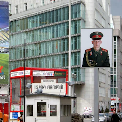 Checkpoint monter märkt "U.S. Army Checkpoint" med ett stort porträtt av en uniformerad soldat i bakgrunden, i ett stadsområde.