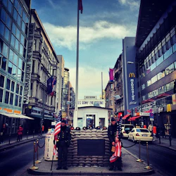 Två vakter står med amerikanska flaggor vid US Army Checkpoint på en livlig stadsgata, omgiven av historiska och moderna byggnader.
