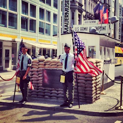 Två män i militära uniformer står med amerikanska flaggor bredvid en kopia av den amerikanska arméns Checkpoint vid Checkpoint Charlie i Berlin.