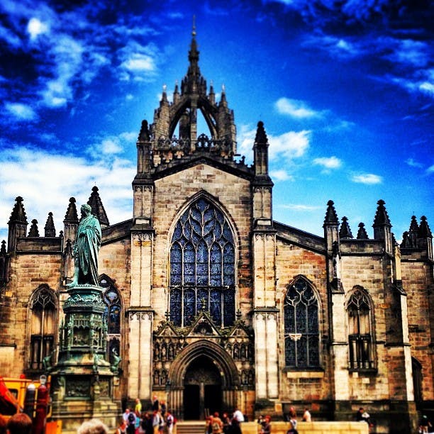 Gotisk katedral med detaljerad arkitektur och ett stort glasmålningsfönster under en vibrerande blå himmel; staty och människor i förgrunden.