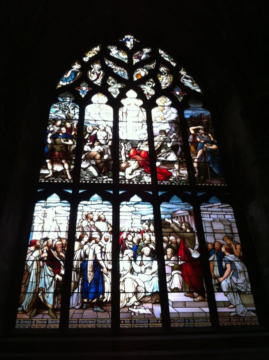 Ett glasmålningsfönster som visar olika historiska och religiösa scener med många figurer i färgglada klädsel och arkitektoniska detaljer.