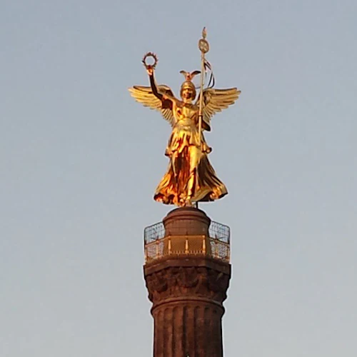 Gyllene bevingad staty med en krans och en stav ovanpå en hög pelare mot en klar himmel.