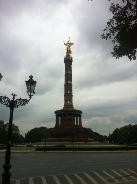 En hög pelare med en gyllene staty på toppen, omgiven av träd, under en molnig himmel, med en gata och utsmyckade lyktstolpar i förgrunden.