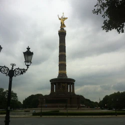 En hög pelare med en gyllene staty på toppen, omgiven av träd, under en molnig himmel, med en gata och utsmyckade lyktstolpar i förgrunden.