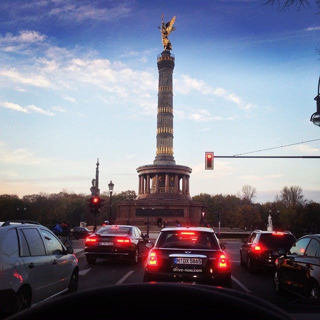 Bilar stannade vid ett trafikljus framför segerkolonnen med en guldstaty ovanpå, under en blå himmel med lätta moln.