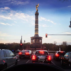 Bilar stannade vid ett trafikljus framför segerkolonnen med en guldstaty ovanpå, under en blå himmel med lätta moln.