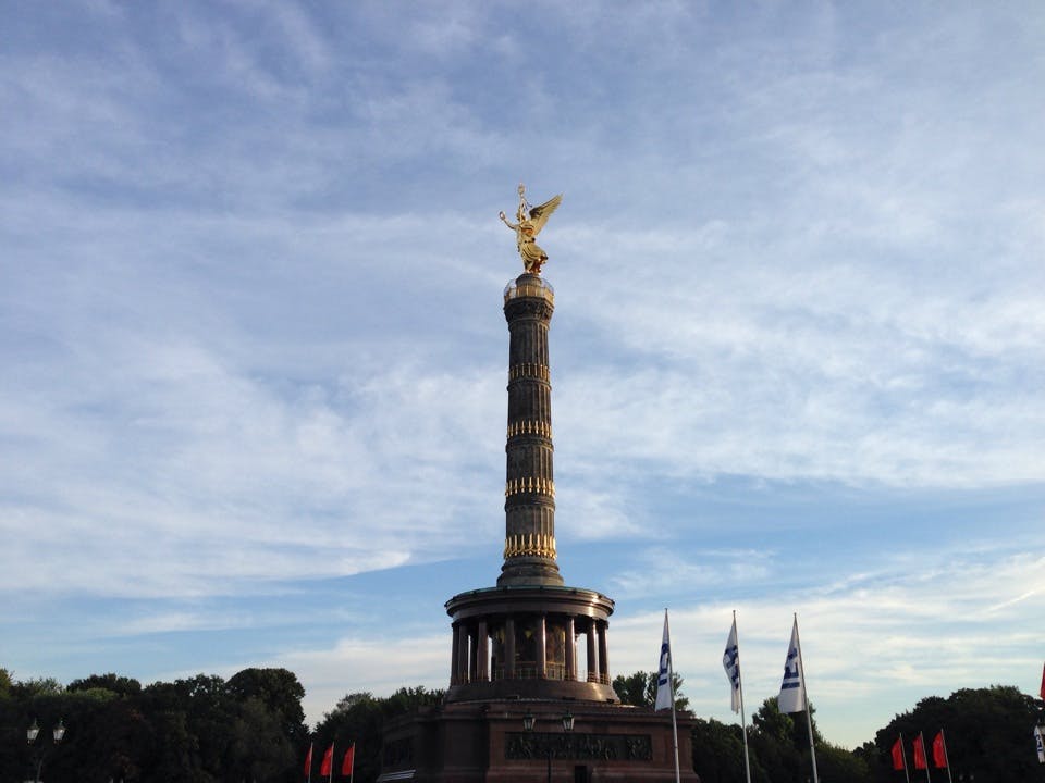 En gyllene staty ovanpå en hög pelare med flaggor och träd nedanför, under en delvis molnig himmel.