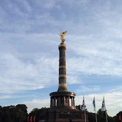 En gyllene staty ovanpå en hög pelare med flaggor och träd nedanför, under en delvis molnig himmel.