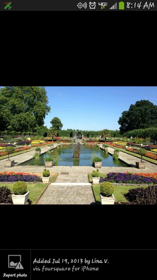 Симметричный сад с центральным отражающим бассейном, красочными клумбами и растениями в горшках. Густые зеленые деревья окружают сад.