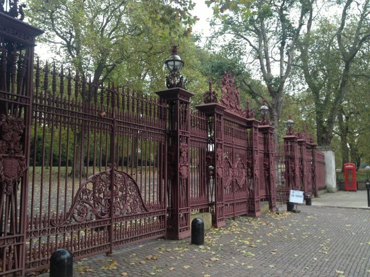 Utsmyckade, höga, röda järnportar med invecklade mönster, i en park med träd och en röd telefonkiosk i bakgrunden.
