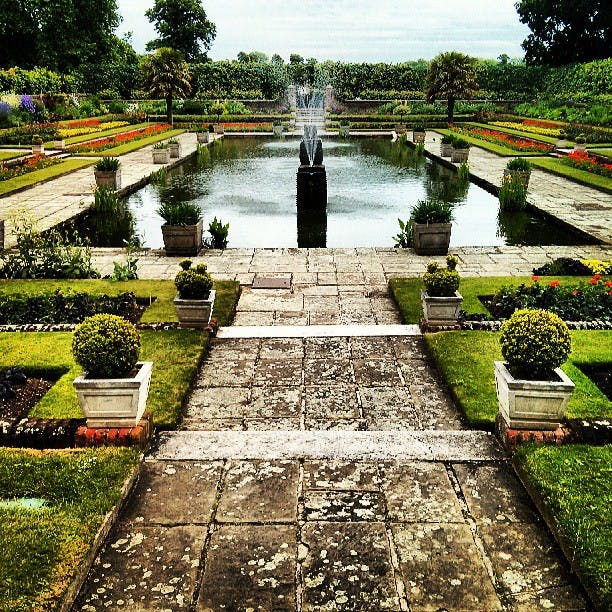 En formell trädgård med en central fontän, symmetriska stigar, välskötta häckar och blomsterrabatter som omger en rektangulär damm.