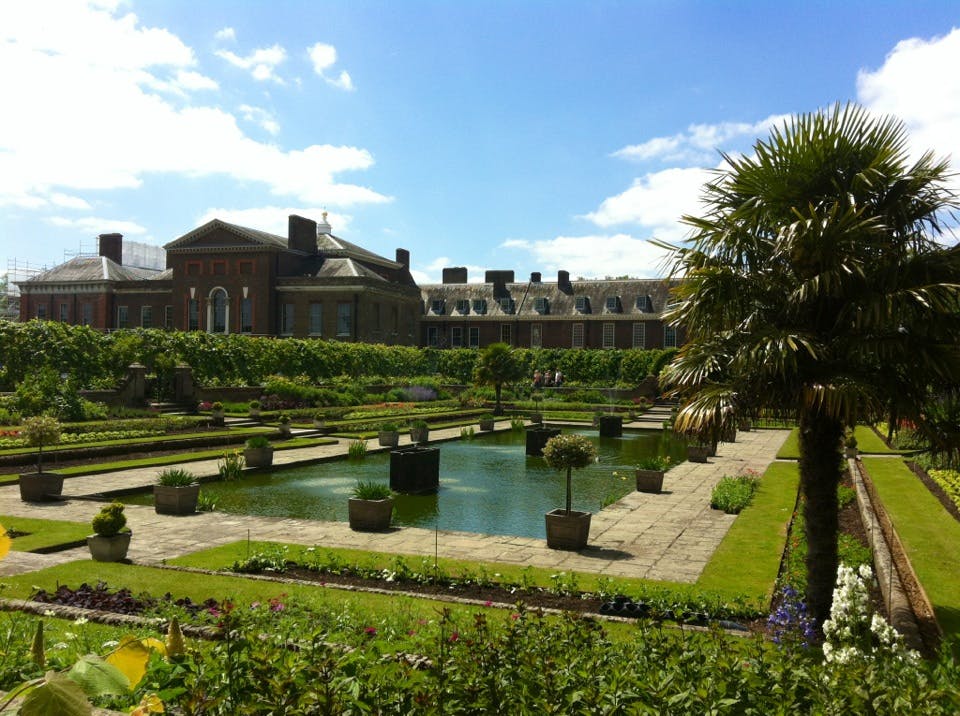 Historisk byggnad med en välskött trädgård, med rektangulära pooler, blomsterrabatter, krukväxter och ett palmträd en solig dag.