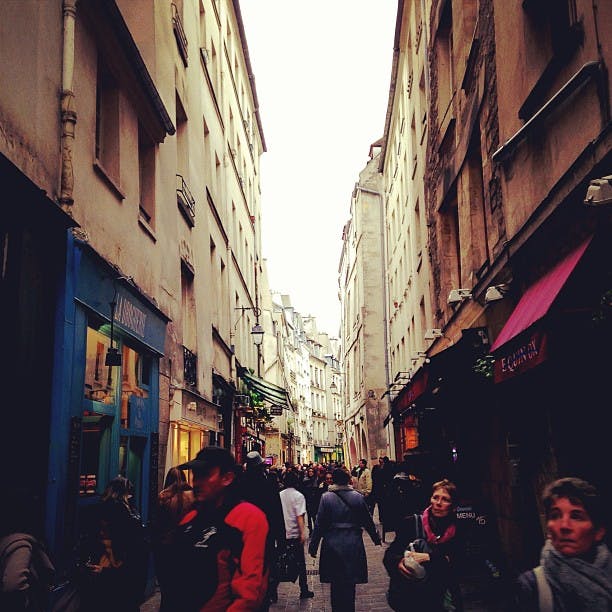 Narrow street with tall, old buildings, filled with pedestrians. Shops on both sides have colorful signs and awnings.