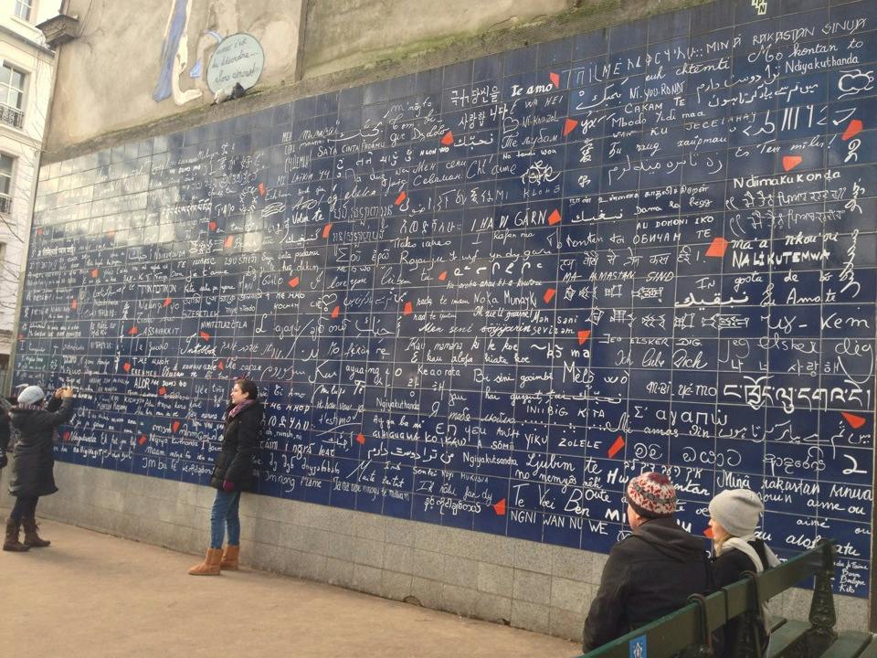 Ludzie oglądający i fotografujący ścianę pokrytą napisem „I love you” w wielu językach w miejscu publicznym.