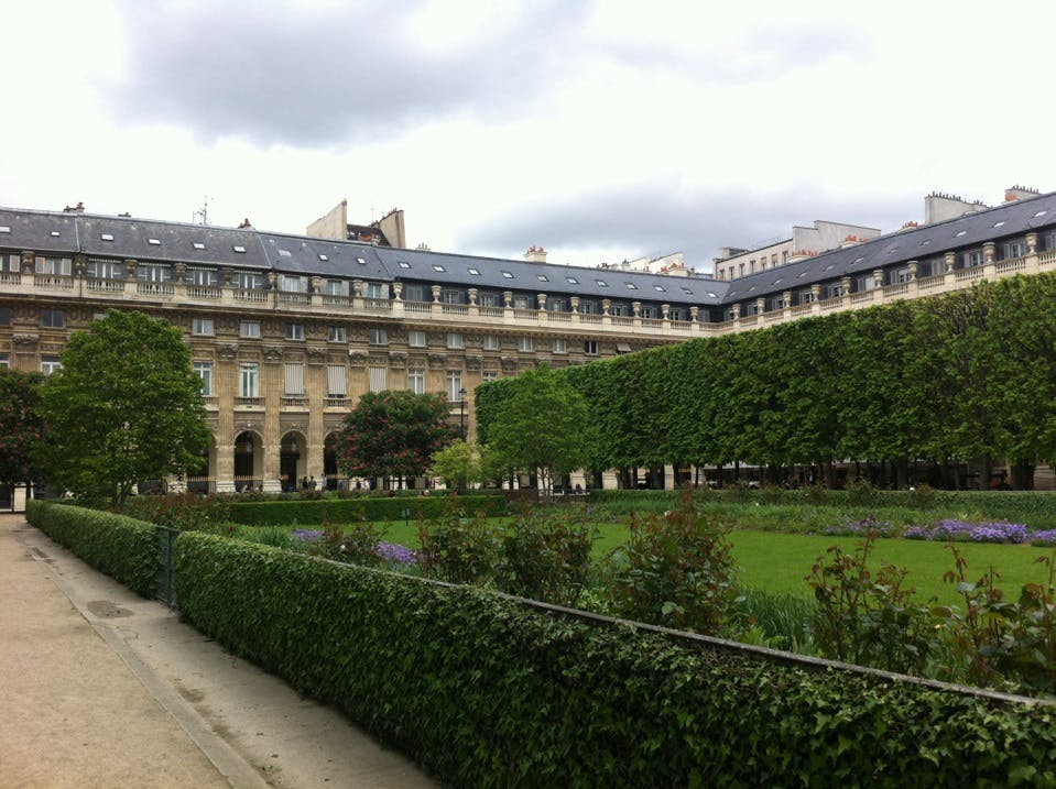 A manicured garden with trimmed hedges and trees, surrounded by a historic building under a cloudy sky.