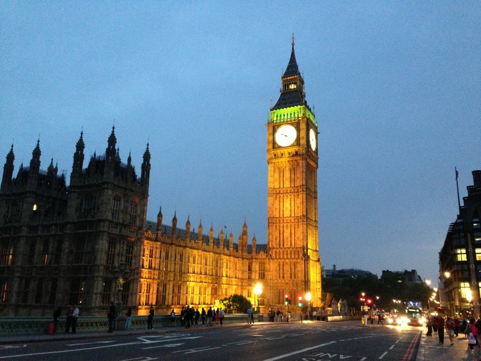 Big Ben klocktorn i skymningen, upplyst med ett grönt och gult ljus. Människor, bussar och bilar på den närliggande gatan.