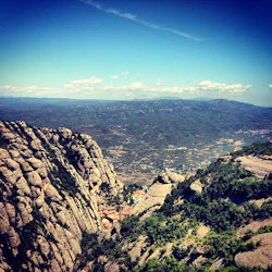 En hög bergsutsikt med steniga klippor, grön vegetation och en liten by i dalen under en blå himmel med spridda moln.