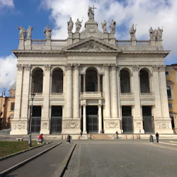 Utsmyckad fasad av en stor byggnad med höga pelare, statyer på taket och människor som går på en stenlagd innergård under en delvis molnig himmel.