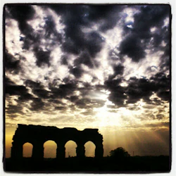 Molnig himmel med solstrålar bakom uråldriga stenbågar siluetterade vid horisonten.