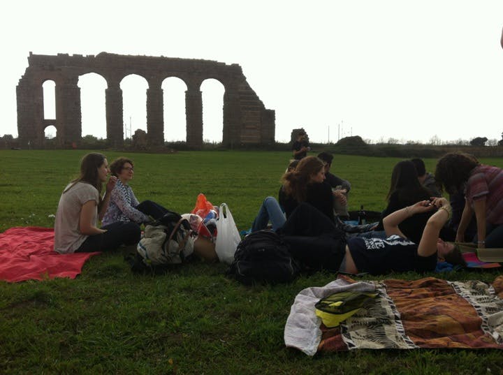 Människor som har en picknick på ett gräsbevuxet fält med ruiner av en gammal akvedukt i bakgrunden.