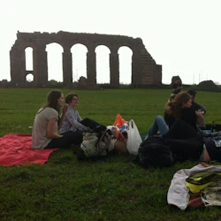 Människor som har en picknick på ett gräsbevuxet fält med ruiner av en gammal akvedukt i bakgrunden.