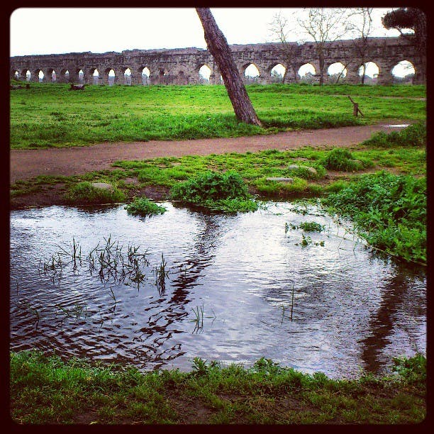 Forntida stenakvedukt med valv, grönt gräs, en smutsväg och en liten damm med reflekterande träd i förgrunden.