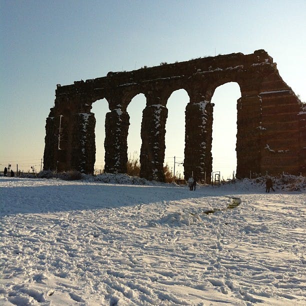 Ancien aqueduc couvert de neige avec de grandes arches, des personnes dispersées en arrière-plan et un ciel dégagé.