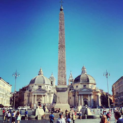En hög obelisk flankerad av två kupolformade byggnader på ett livligt torg med människor som går och sitter under en klarblå himmel.