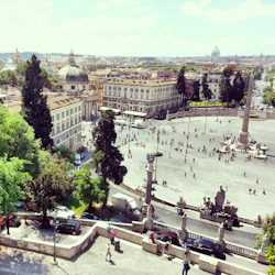 En flygbild av ett stort torg med en hög obelisk, omgiven av historiska byggnader, träd och många människor som går.