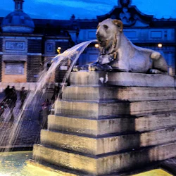 Lejonstatyfontän med vatten som strömmar nerför trappor, mot en utsmyckad byggnad i skymningen.
