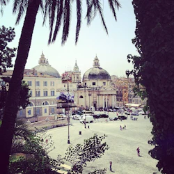 Ett stort torg med en historisk byggnad med kupoler och torn, inramad av palmer och grönska, med människor som går runt.