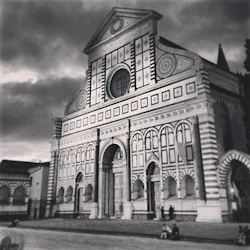 Svartvit bild av en byggnadsfasad i gotisk stil med välvda fönster och cirkulära mönster; några personer sitter eller står i närheten.