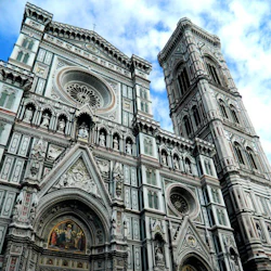 Utsmyckad katedralfasad med detaljerade sniderier, statyer, runda fönster och ett högt klocktorn mot en delvis molnig himmel.