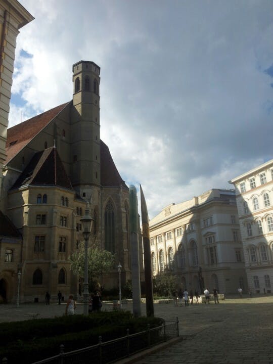Una chiesa in stile gotico e gli edifici circostanti sotto un cielo parzialmente nuvoloso con persone che camminano in un cortile.