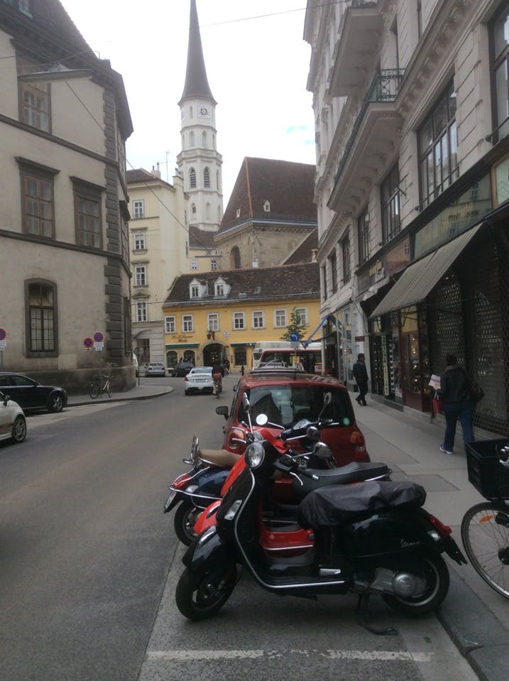 Gatuscen med parkerade skotrar i förgrunden, bilar, fotgängare och en kyrka med en hög spiral i bakgrunden.
