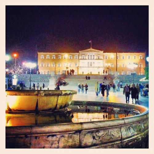 Fontän på ett torg på natten, med en stor upplyst byggnad och människor som går i närheten.