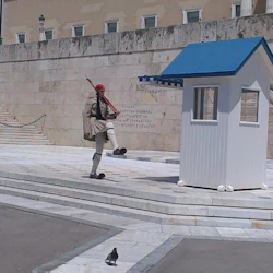 En vakt i traditionell uniform utför en marsch bredvid ett vitt och blått vakthus framför en historisk byggnad. En duva ligger på trottoaren.