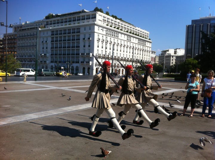 Marcherande vakter med gevär i traditionella uniformer och röda hattar, omgivna av duvor och turister på ett torg. Stor byggnad i bakgrunden.