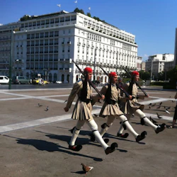Marcherande vakter med gevär i traditionella uniformer och röda hattar, omgivna av duvor och turister på ett torg. Stor byggnad i bakgrunden.